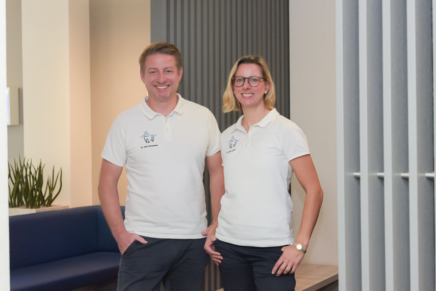 Zwei Zahnärzte in weißen Poloshirts stehen in einer modernen Praxis mit einer blauen Couch und Pflanzen im Hintergrund. Sie lächeln freundlich in die Kamera.