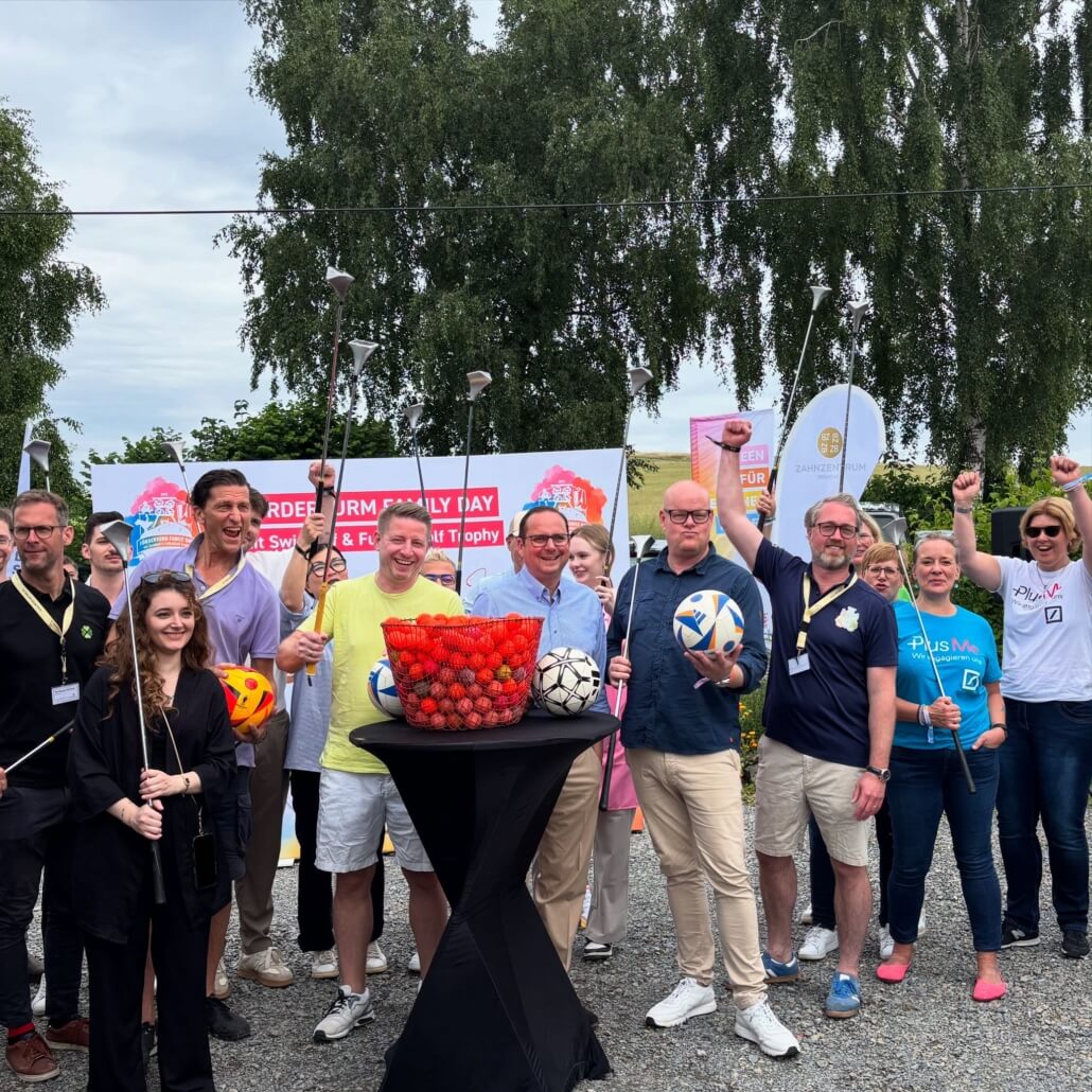 Eine Gruppe von Menschen steht zusammen und hält verschiedene Fußbälle und Golfschläger. Im Vordergrund steht ein Tisch mit einer Schüssel voller roter Bälle. Im Hintergrund sind Banner zu sehen.