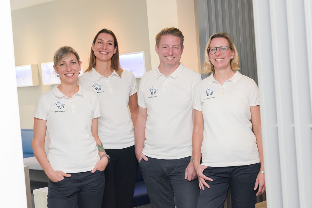 Ein Team von vier Personen in weißen Poloshirts steht lächelnd in einem modernen Raum. Sie wirken freundlich und professionell, bereit, Patienten zu empfangen.