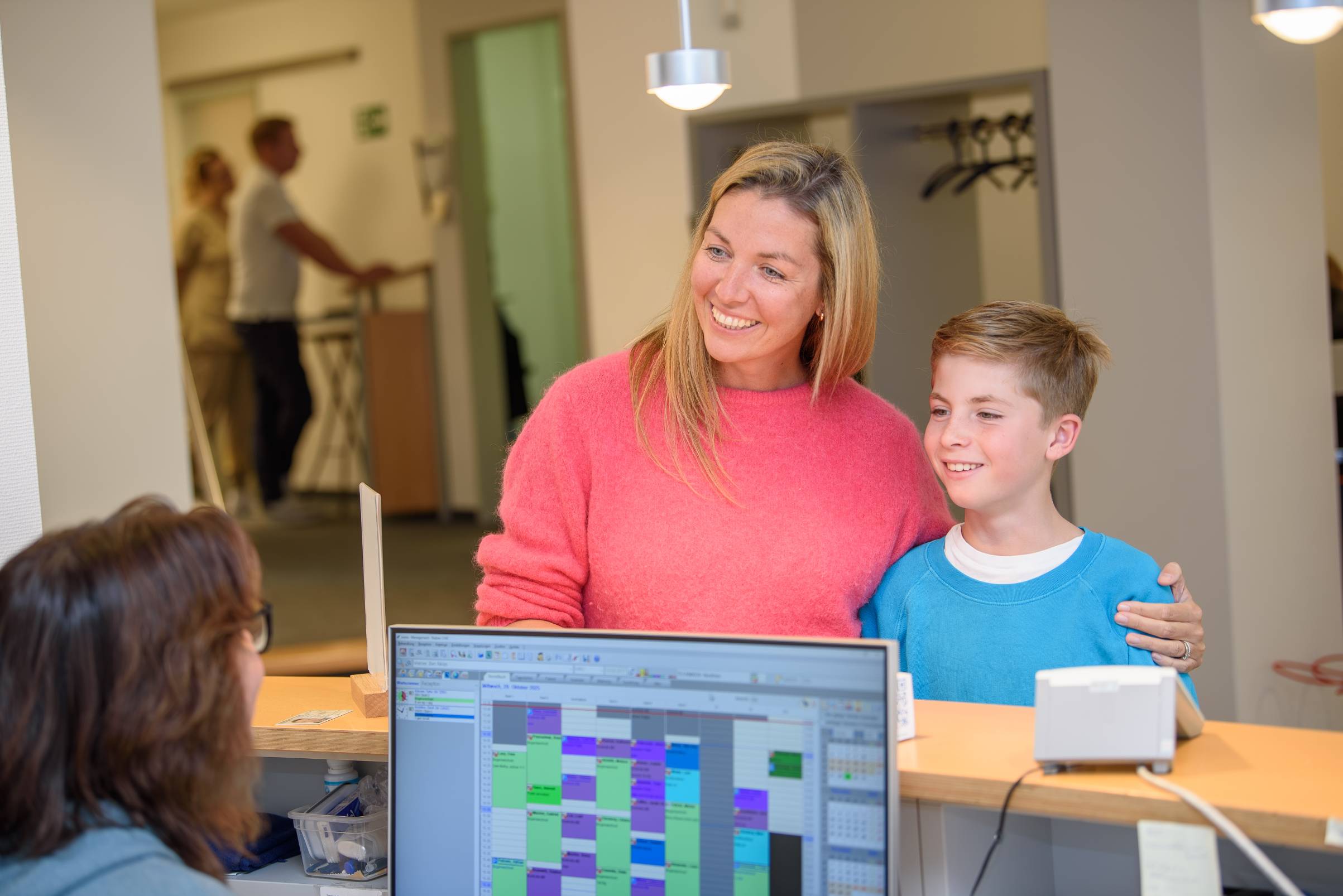 Eine lächelnde Mutter in einem pinken Pullover steht mit ihrem Sohn in einem blauen Pullover an einem Empfangstresen, während eine Mitarbeiterin am Computer arbeitet.